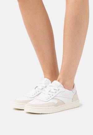 Piernas con zapatillas bajas blancas con detalles en ante beige y cordones blancos, de pie sobre un fondo claro y sencillo.