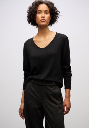 Schwarzer V-Ausschnitt-Pullover aus weichem Material, kombiniert mit dunklen, maßgeschneiderten Hosen mit Seitentaschen und Knopfdetails.