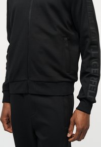 Κοντινό πλάνο ενός ατόμου που φοράει μαύρο μπουφάν με φερμουάρ και αντίστοιχο παντελόνι με ένα αχνό κάθετο λογότυπο στο μανίκι.