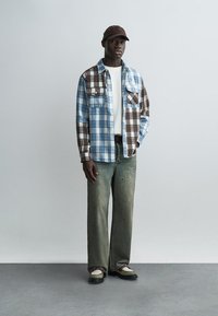 Muž nosící hnědou čepici, modro-hnědou kostkovanou košili přes bílé tričko, vybledlé zelené široké džíny a béžovo-černé boty stojící u šedé zdi.