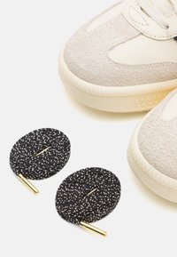 Sneaker con una parte superiore in suede beige e suola testurizzata bianco sporco, accompagnate da due lacci a pois neri e bianchi con punte dorate.