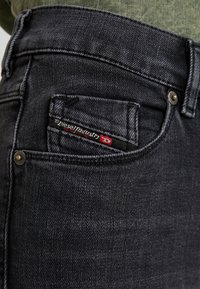 Primer plano del bolsillo de unos vaqueros de mezclilla negros con la etiqueta de Diesel Industry y remaches metálicos, sobre una persona que lleva una camisa verde oliva.