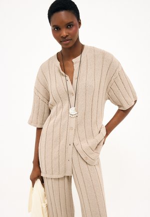 Žena oblečená v béžovom rebrovanom krátkormenom zapínacom tričku na gombíky a zodpovedajúcich nohaviciach, držiaca v jednej ruke svetlú látku.