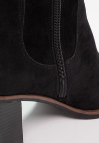 Botte de cheville en suede noir avec une fermeture éclair sur le côté, un talon bloc empilé et des bordures marron le long du bord de la semelle sur un fond blanc.