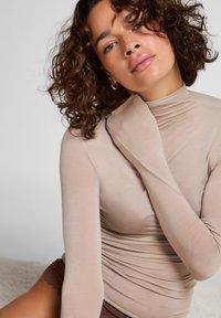 Beige langekleurige top met lange mouwen en een hoge nek, gemaakt van zachte, rekbare stof. Model heeft krullend haar en draagt bruine kantenshorts.