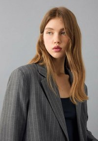 Grijze blazer met krijtstrepen, een ontspannen pasvorm, inkeping revers en een zachte stof. Draagt over een zwart topje, passend bij een neutrale achtergrond.