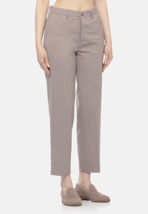 Donna che indossa pantaloni grigi su misura alla caviglia e mocassini intrecciati abbinati senza lacci, in piedi davanti a uno sfondo chiaro uniforme.
