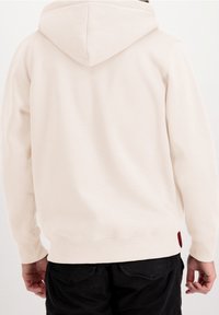 Lichtbeige hoodie gemaakt van zachte stof, met een rugnaad, geribde boorden en een klein rood accent aan de onderzijde.