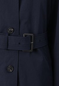 Marine belted kåpe med knappestenging, teksturert stoff og glatt overflate, bred belte og enkel svart spenne.