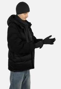 Black puffer jacket with a hood, paired with a gray scarf and gloves. The individual is adjusting the gloves, wearing a dark knit beanie.