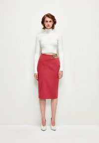 Top à manches longues blanc à nervures associé à une jupe rouge ajustée arrivant au genou avec un ourlet asymétrique et une ceinture en accent doré. Talons couleur crème.