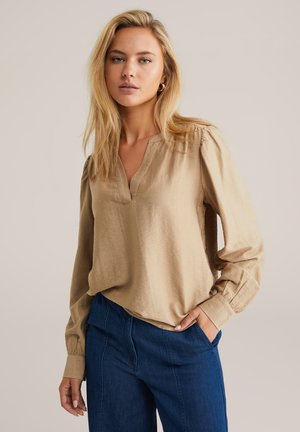 Blouse beige à manches longues avec un col en V, épaules froncées et coupe décontractée, associée à un pantalon en denim bleu foncé. Texture lisse.
