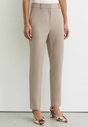 Beežid kohandatud püksid vööaasadega, mida kannab inimene, kombineeritud pruunide ja valgete lehmakujuliste kõrge kontsaga kingadega.