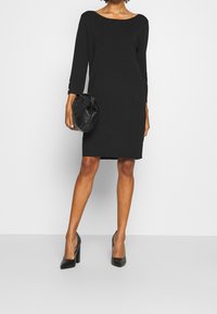 Robe noire longueur genou avec un décolleté bateau, des manches trois-quarts et des détails en boutons métalliques. Assortie avec des escarpins noirs pointus et un sac noir.