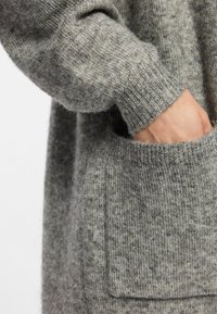 Suéter gris de punto con una textura suave, que cuenta con un bolsillo en la parte frontal y puños acanalados. El material parece cálido y acogedor.