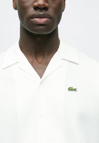 Biela polokošeľa s krátkym rukávom a textúrovaným vzorom, má klasický golier a vyšívané zelené logo krokodíla na ľavej strane hrudníka.