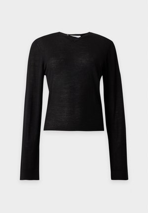 Frau steht mit den Händen in den Taschen, trägt ein schwarzes langärmeliges Oberteil, weite braune Hosen und schwarze spitze Schuhe.