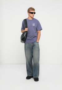 Camiseta de algodón morada con gráfico blanco, pantalones de denim azul holgados, gafas de sol negras y un bolso de hombro de cuero negro. Fondo liso.