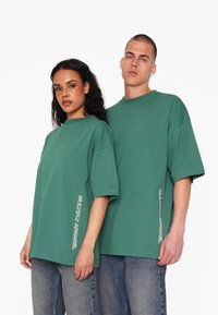 T-shirt oversized verde in cotone con maniche corte, caratterizzato dal testo "MULTIPLY APPAREL" stampato verticalmente sul lato sinistro.