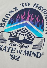 Tricou grafic cu un design cu flacăra în roz și albastru, cu accente în pattern tablă de șah. Textul spune "BRONX CĂTRE BROOKLYN, New York, '92."