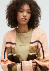Multicolored knit cardigan featuring pink, brown, and navy stripes. Soft texture with oversized sleeves and a relaxed fit over a beige t-shirt.