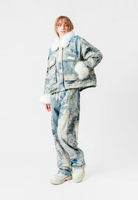 Chaqueta de patrón azul y blanco con cuello de piel sintética y múltiples bolsillos, combinada con pantalones holgados a juego y calzado de color claro.