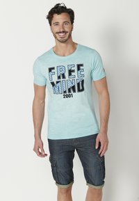 Világoskék, rövid ujjú póló, amelyen vastag fekete és kék betűkkel a "FREE MIND 2001" felirat látható, sötét farmer sorttal párosítva.