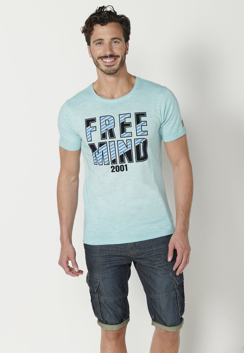 Világoskék, rövid ujjú póló, amelyen vastag fekete és kék betűkkel a "FREE MIND 2001" felirat látható, sötét farmer sorttal párosítva.