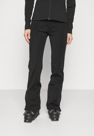 Persoon die zwarte, nauwsluitende skibroek, zwart jack met ritssluiting en zwarte skischoenen draagt, staand tegen een effen witte achtergrond.