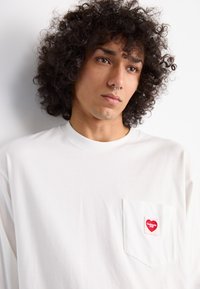 White long-sleeve T-shirt with a front pocket featuring a red heart logo. Soft fabric with a relaxed fit and round neckline.