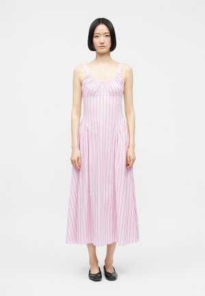 Γυναίκα με ίσια μαύρα μαλλιά, φορώντας ένα αμάνικο, ροζ φόρεμα μέχρι τους αστραγάλους με κόκκινες κάθετες ρίγες, στέκεται μπροστά σε λευκό φόντο.