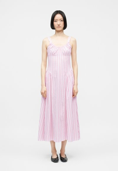 Frau mit glattem schwarzem Haar, die ein ärmelloses, bodenlanges rosa Kleid mit roten vertikalen Streifen trägt und vor einem schlichten weißen Hintergrund steht.