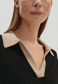 Maglione nero con colletto beige, presenta un design a V. Il materiale sembra soffice con una texture liscia. Dettaglio di cucitura sottile visibile.