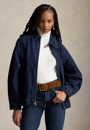 Mujer con el pelo largo trenzado que lleva una chaqueta vaquera azul oscuro sobre un jersey de cuello alto blanco y vaqueros azules con un cinturón marrón, sobre un fondo neutro.