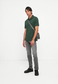 Groene katoenen polo met kraag, grijze denim jeans en zwarte schoenen. Kleine logodetail op het shirt. Schoudertas in het grijs.