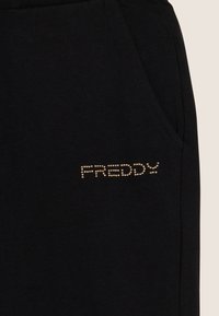 Pantalones de chándal negros de algodón con bolsillos laterales y un logo dorado con tachuelas que dice "FREDDY" en el muslo izquierdo. Textura suave.
