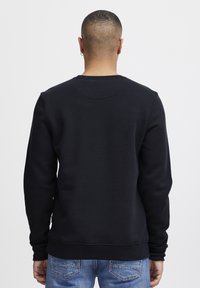 Felpa nera con collo a giro, polsini e orlo a costine; tessuto misto cotone; indossata con jeans blu, vista da dietro.