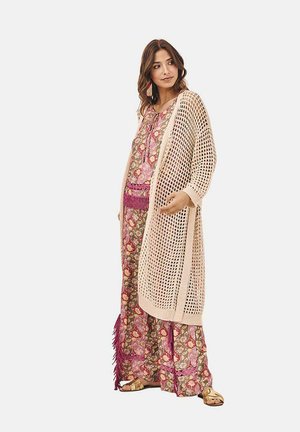Cárdigan de punto y ganchillo en crema, superpuesto sobre un vestido maxi con estampado floral en tonos rosa, verde y púrpura, adornado con flecos.