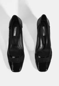 Schwarze High-Heels mit glänzender Lackspitze und einem obermaterial, das an Mesh erinnert. Viereckige Zehenform und glatte Innenauskleidung. Markenlabel sichtbar.