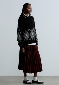 Maglione nero con motivo a diamante grigio, abbinato a una gonna plissettata color bordeaux. La modella indossa calzini bianchi e scarpe nere. Sfondo semplice.