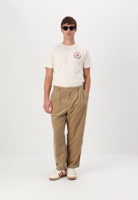 Camiseta de algodón beige con gráfico floral rojo, combinada con pantalones de pana caqui. El modelo lleva gafas de sol negras y zapatillas blancas con acentos rojos.
