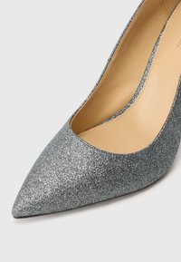 Silver glitter high-heeled shoe with a pointed toe and a cutout design. Smooth beige insole and subtle stitching along the edges.