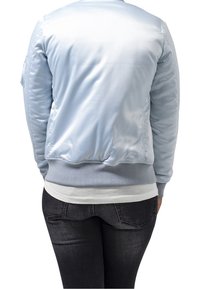 Jasnoniebieska satynowa kurtka bomber z prążkowanymi mankietami i ściągaczem, z tylną kieszenią i gładką teksturą. Nosi się ją na białą koszulę i ciemne dżinsy.