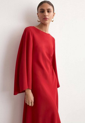 Frau mit zurückgekämmtem Haar, die goldene Creolen trägt und ein rotes Kleid mit langen, weiten Ärmeln, stehend vor einer hellen Wand.