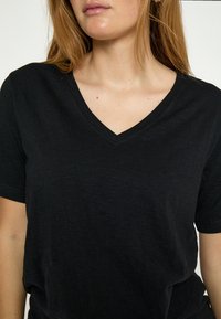 Vrouw draagt een effen zwart T-shirt met korte mouwen en V-hals, waarbij de nek, schouders en bovenkant van de borst met een lichte huid en lang haar zichtbaar zijn.