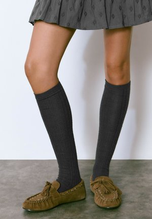 Calcetines gris oscuro hasta la rodilla con textura acanalada, combinados con unos zapatos mocasín de ante marrón que presentan acentos de flecos y tachuelas metálicas.