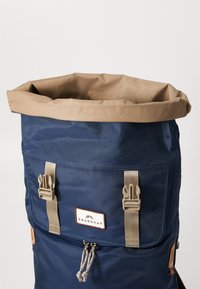 Navy blå rulletopp ryggsekk med beige tekstiltrim, justerbare stropper, en frontlomme og en unik snøring.