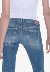 Jeans de mezclilla en azul claro con una cintura de altura media, cinco bolsillos y un parche de cuero en la parte posterior. La tela tiene una textura suave y desgastada.