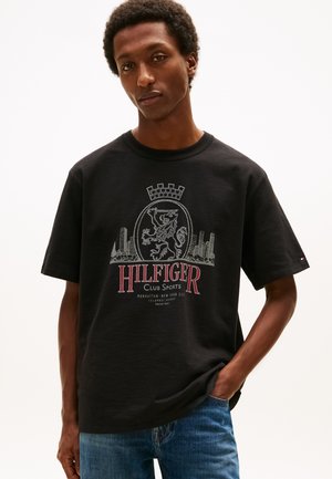 Mężczyzna noszący czarną koszulkę Hilfiger Club Sports z herbem lwa i grafiką panoramy miasta, w połączeniu z niebieskimi dżinsami, stojący na białym tle.