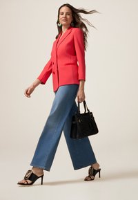 Blazer corallo, jeans a gamba larga blu, borsa nera e sandali neri con tacco e chiusura decorativa. Il modello indossa orecchini pendenti e ha i capelli fluenti.